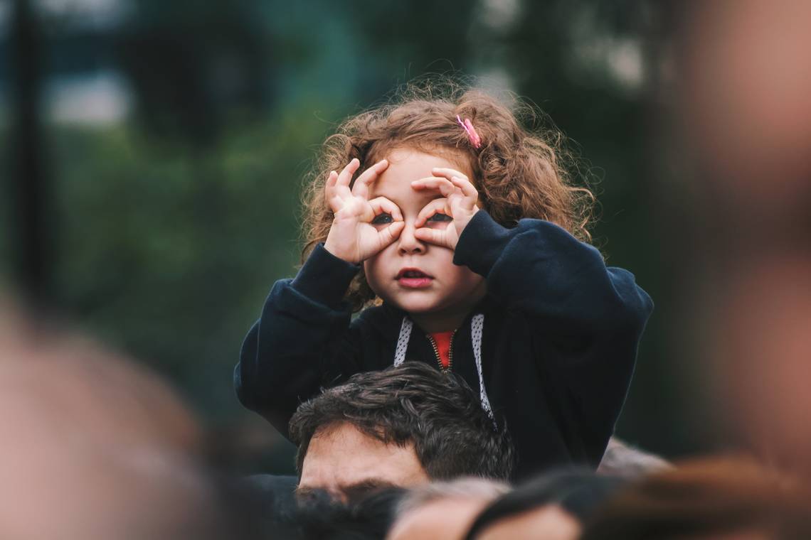Ein rothaariges Mädchen sitzt auf den Schultern eines Mannes und schaut durch ihre Brille, die sie mit Daumen und Zeigefinger formt. | © Edi Libedinsky via Unsplash